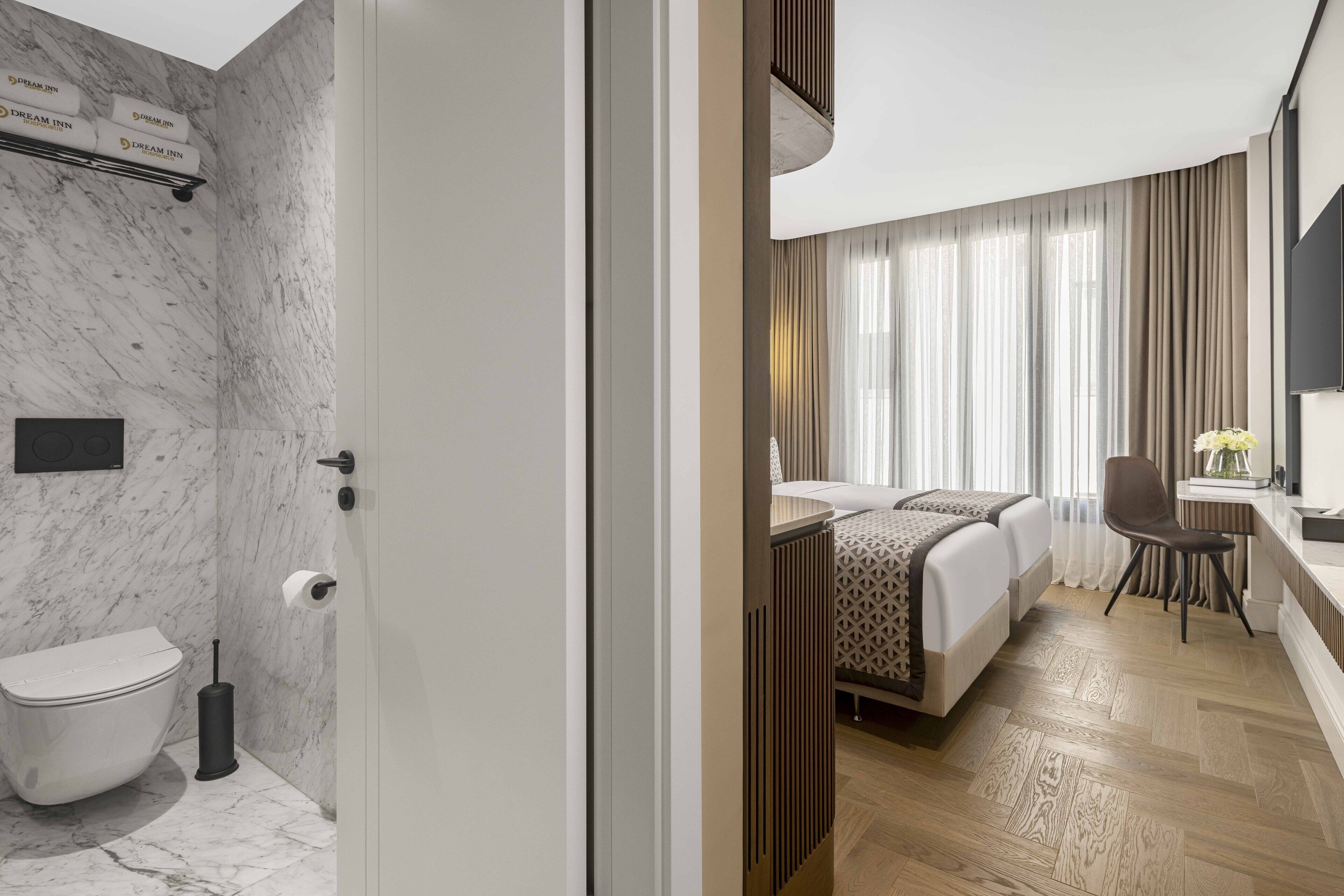Une chambre d’hôtel moderne avec une salle de bain en marbre, un lit recouvert d’une courtepointe à motifs, un plancher en bois et une grande fenêtre avec des rideaux.
