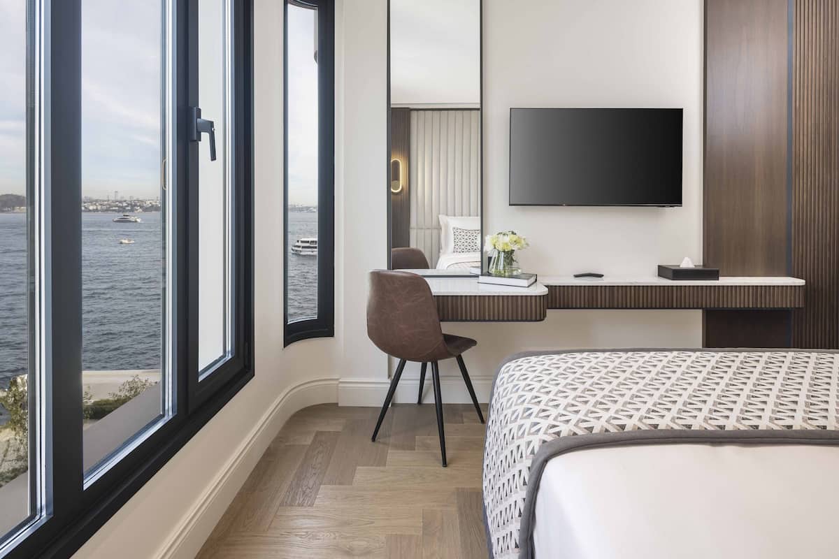 Ein Hotelzimmer mit großem Fenster und Blick auf das Wasser, einem Bett, einem Schreibtisch mit Stuhl und einem Wand-Fernseher.