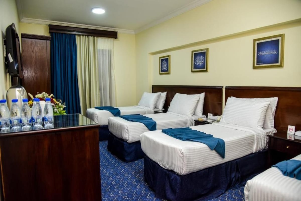 A hotel room with three beds, a desk with water bottles, and framed calligraphy on the wall.