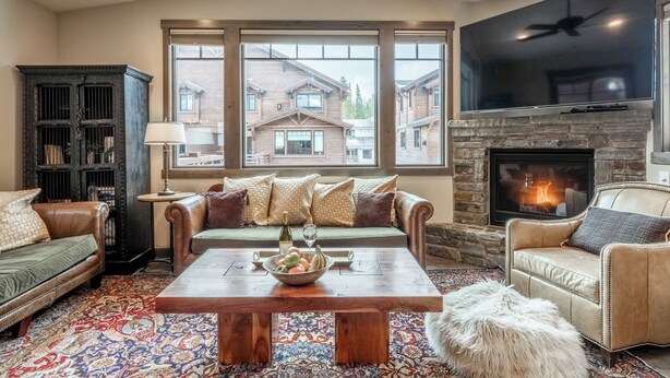 A cozy living room with a fireplace, leather sofas, a coffee table, and a large window offering a view of houses.