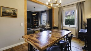 A dining area with a wooden table, chairs, and a chandelier. There is a sofa and a bookshelf in the background.
