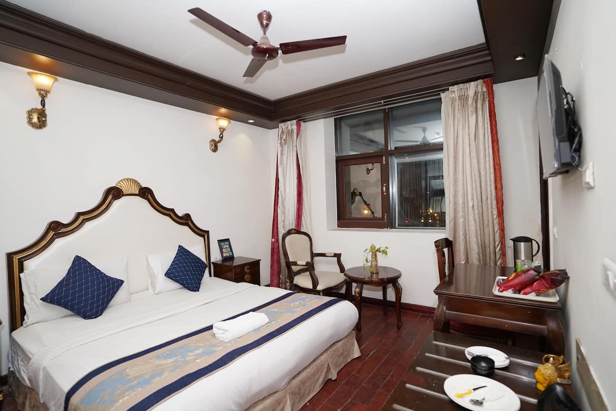 Ein Hotelzimmer mit Bett, Sessel, kleinem Tisch mit Pflanze, Fenster mit Vorhängen und Deckenventilator.