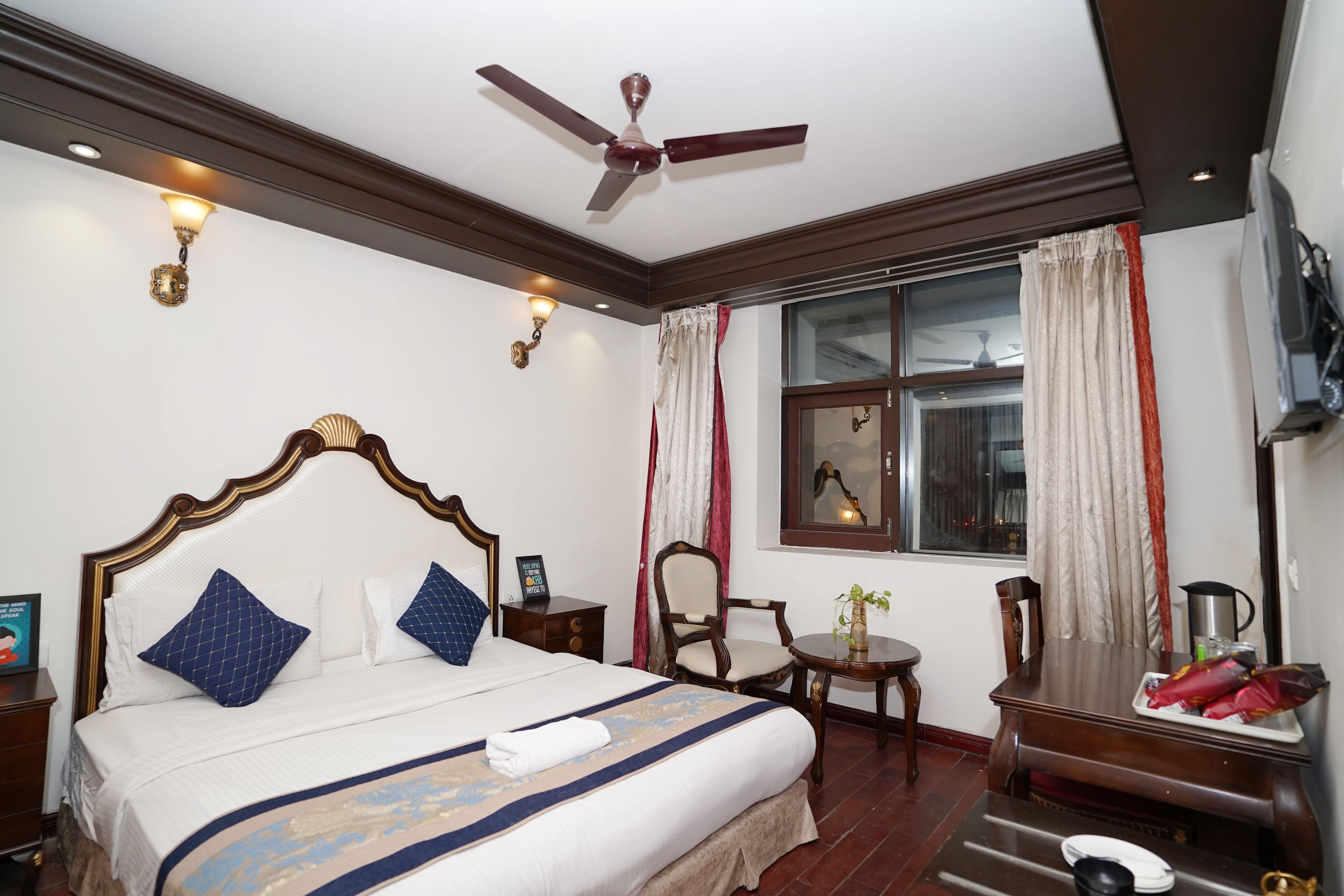 A hotel room with a bed, a ceiling fan, a window with curtains, a small table with a plant, and a TV mounted on the wall.