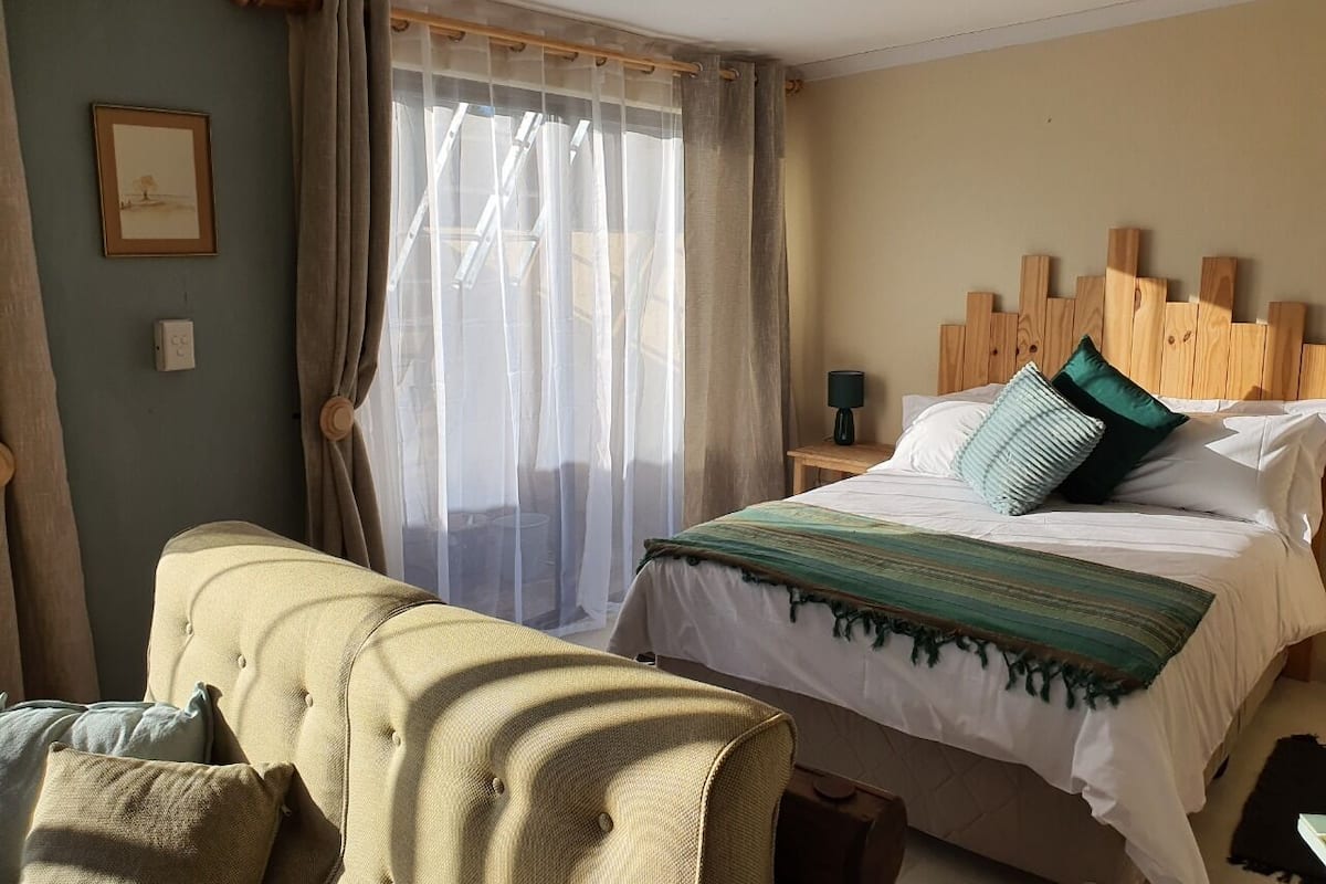 A hotel room with a bed, a sofa, a wooden headboard, a lamp, and a window with sheer curtains.