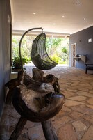 Una silla colgante, una escultura de madera y un suelo de piedra en una habitación con vistas a zonas verdes.