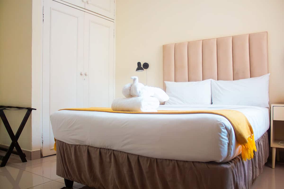 Uma cama bem arrumada com lençóis brancos e saia marrom, um abajur e um guarda-roupa branco.