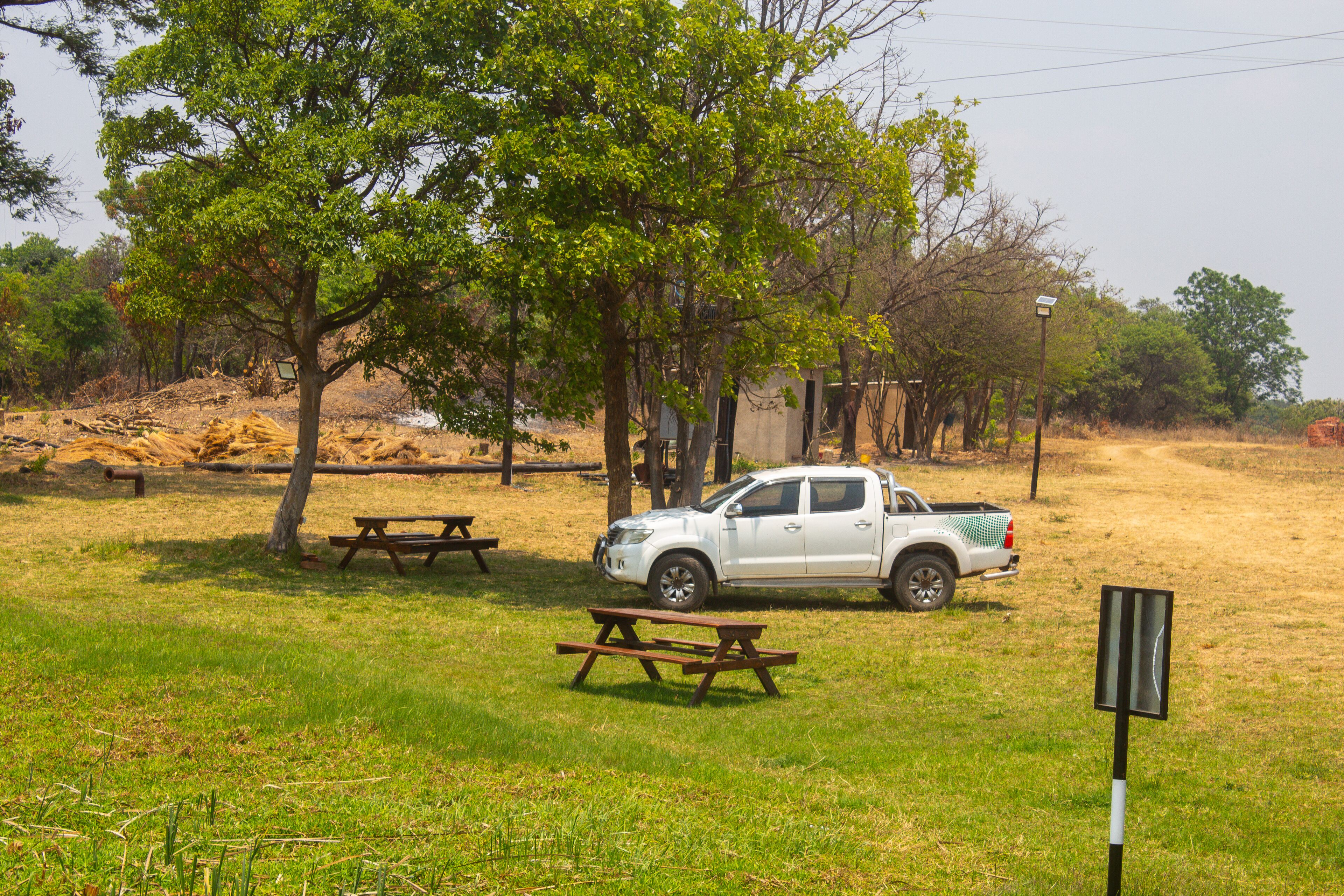 Una camioneta pickup blanca estacionada cerca de mesas de picnic en un área con césped, árboles y un sendero de tierra.