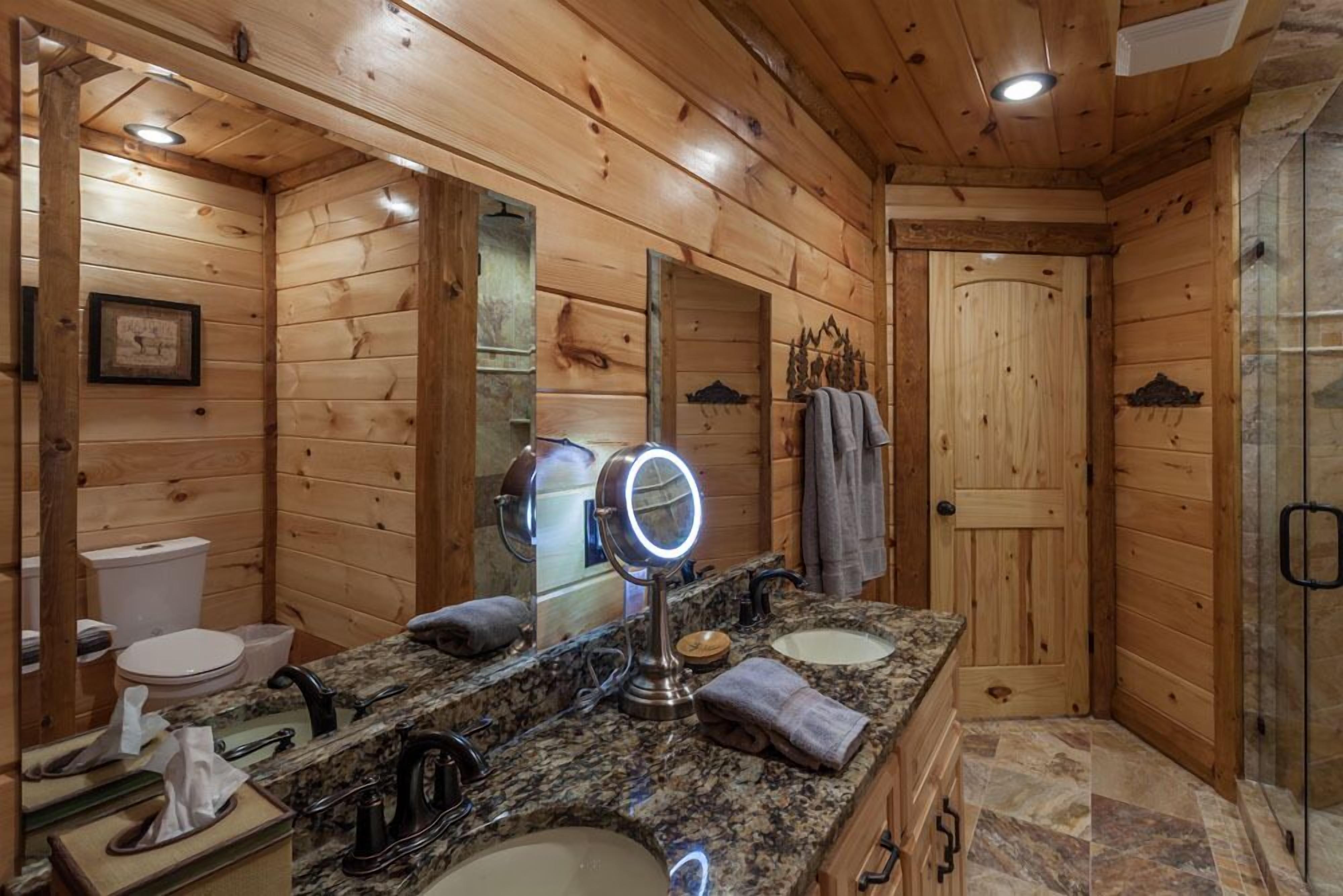 Baño de cabaña de madera con encimera de granito, dos lavamanos, un espejo con iluminación, un inodoro y una zona de ducha.
