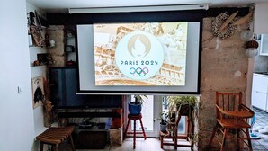 A living room with a large screen displaying the Paris 2024 Olympics logo, a wooden bench, a small table, and a potted plant. - ღ LE JARDIN PARISIEN - quiet with terrace wifi, video projector (Paris)