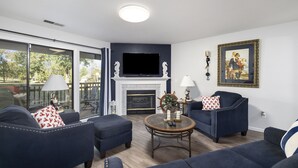 Ein Wohnzimmer mit Kamin, Fernseher und einem großen Fenster mit Blick auf eine Terrasse.