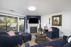 A living room with a fireplace, a television, and a large window overlooking a deck.