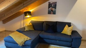 A blue sectional sofa with yellow and dark blue cushions, a yellow throw blanket, and a black floor lamp in a room with wooden beams on the ceiling. - Schöne Ferienwohnung "hubertus" im Dachgeschoss (Holzkirchen)