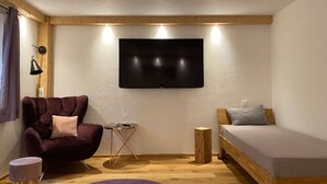 A modern room with a flat-screen TV mounted on the wall, a dark-colored sofa, a small round table, a wooden stool, and a bed with a wooden frame. - Neurenovierte 60m2 Fewo Xaverl zw. Stadt&bergen (Holzkirchen)