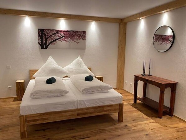 Un lit double avec du linge de lit blanc et deux oreillers, une table de chevet en bois, une table en bois avec des bougies et un tableau représentant un arbre accroché au mur.