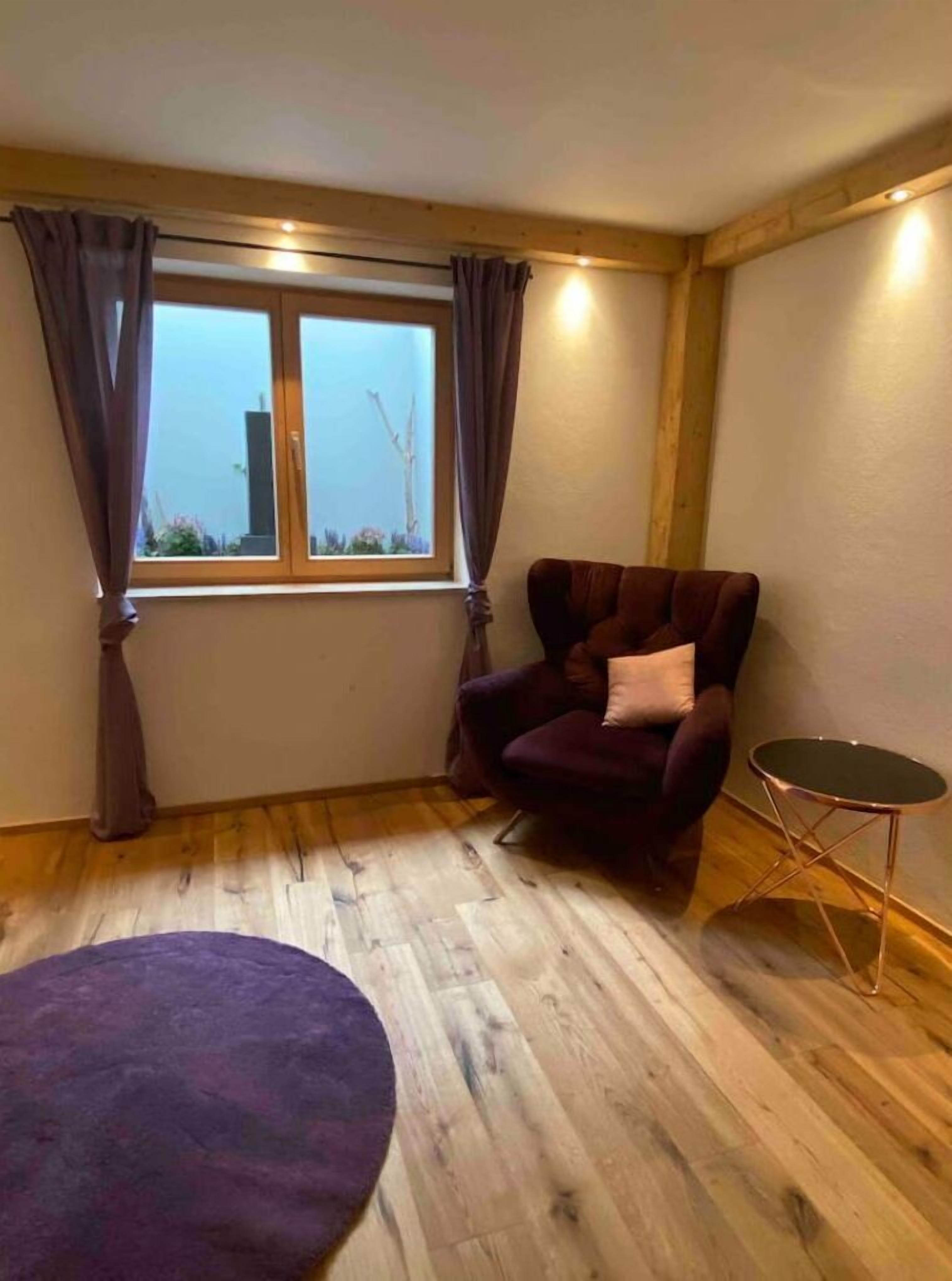 A room with a wooden floor, a purple armchair with a pillow, a small round table, and a window with curtains.