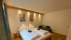 Una cama doble con ropa de cama blanca, un sofá oscuro y dos lámparas fijadas a la pared.
