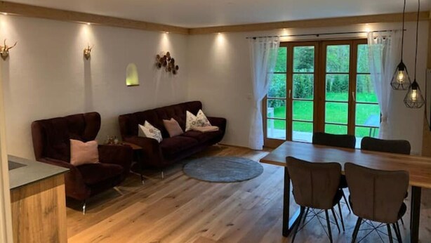 Un salón amplio con suelo de madera, un conjunto de sofá marrón oscuro, una mesa de comedor con cuatro sillas y un ventanal con vistas a zonas verdes.