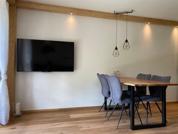 Un comedor con una mesa y sillas de madera, un televisor de pantalla plana en la pared e iluminación colgante.