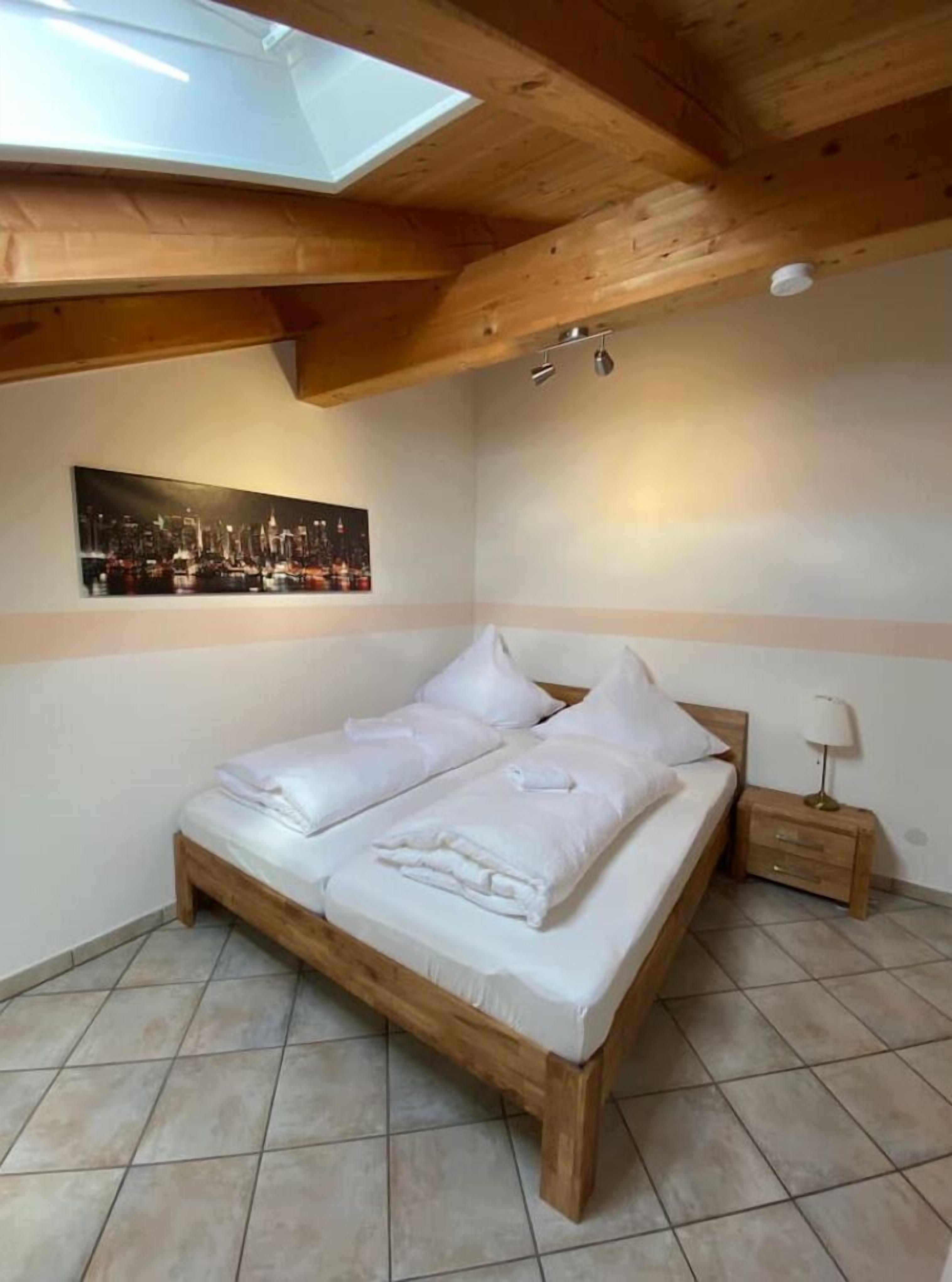 A bedroom with a wooden bed, white bedding, a nightstand with a lamp, a cityscape painting on the wall, and a skylight above.