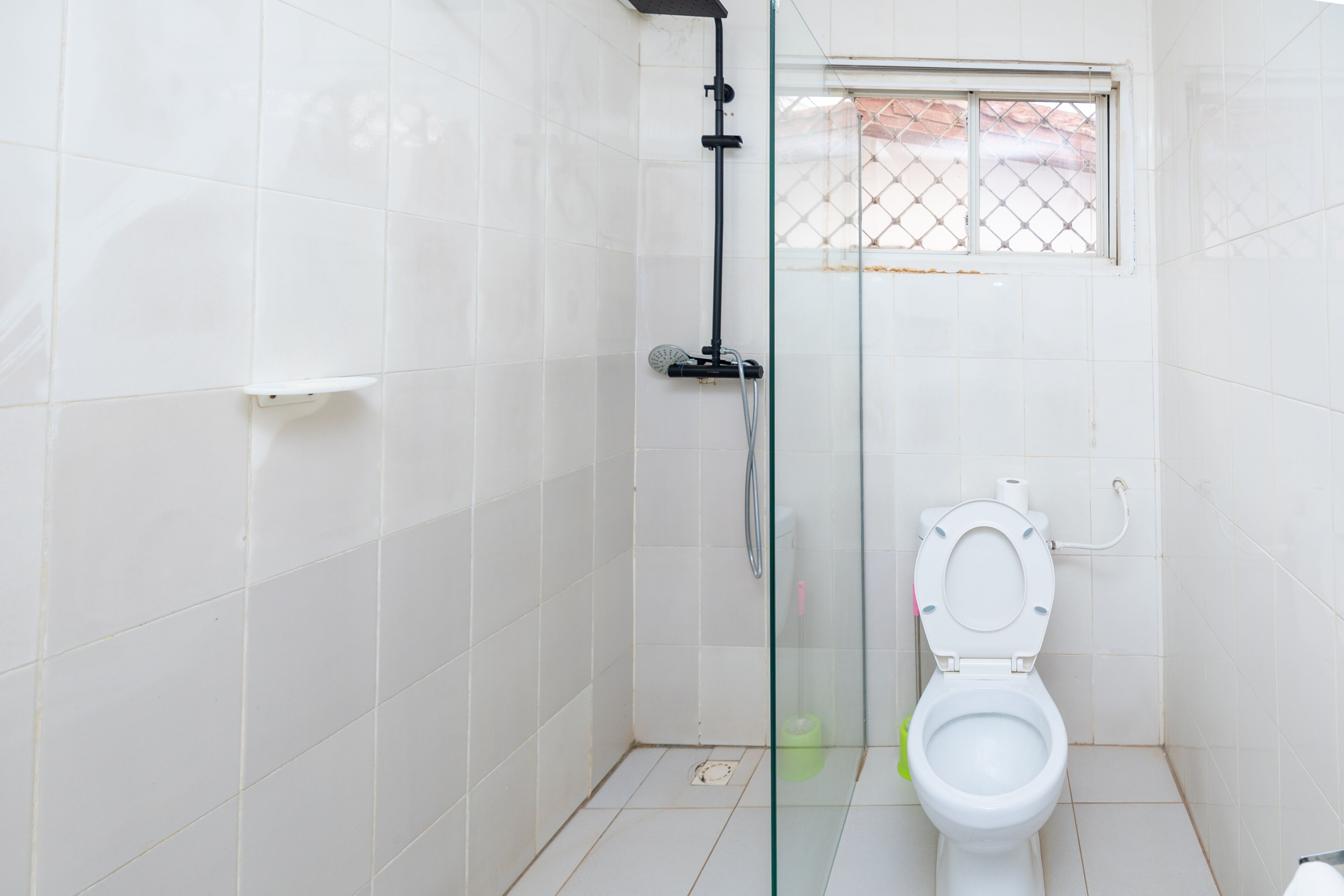 Baño con azulejos blancos, cabina de ducha de vidrio, inodoro y ventana con celosía de metal.