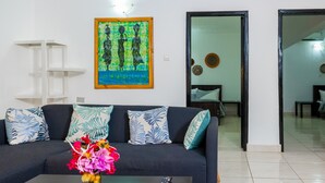 A living room with a dark sofa, patterned cushions, a coffee table with a vase of flowers, and a framed artwork on the wall. - Sarge's Hotel (Serrekunda)
