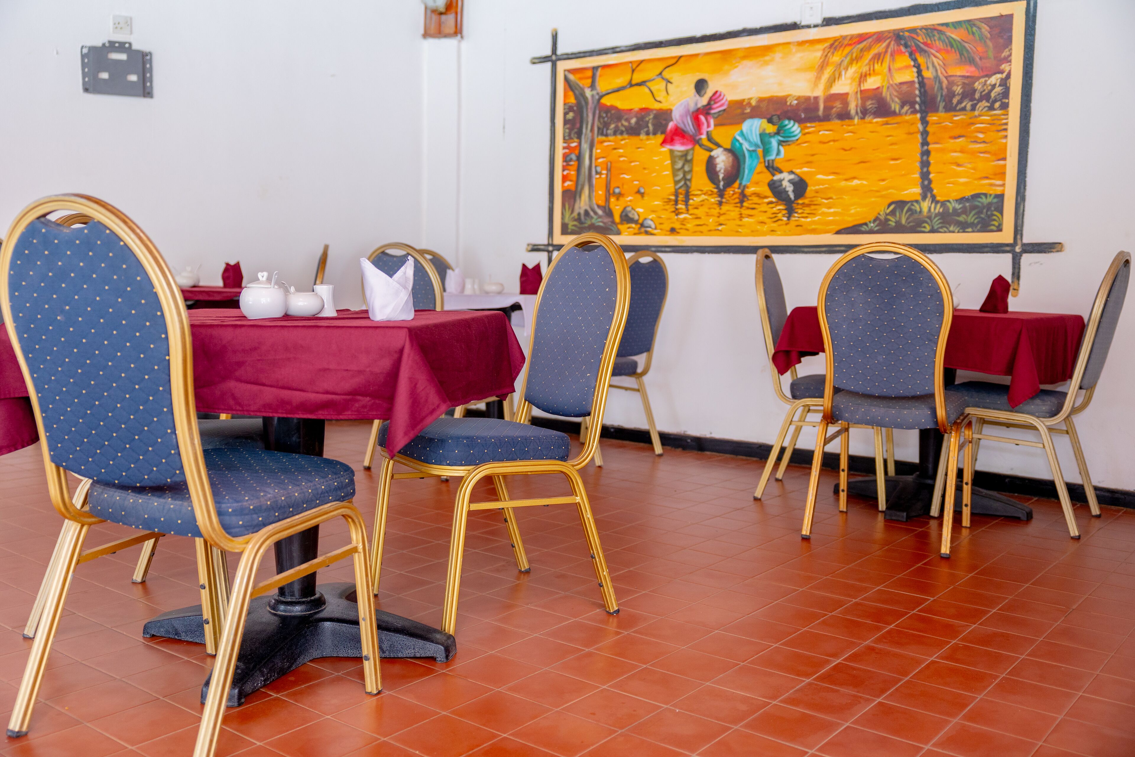 Un coin repas avec une nappe rouge, des chaises bleues à l’armature dorée et un tableau accroché au mur.