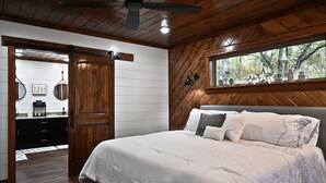 A bedroom with a bed, wooden ceiling, and a window with a view of trees.