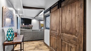 A hallway with a wooden floor, a grey sofa, a TV mounted on a stone wall, and a wooden door.