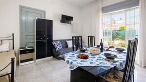 Een eetgedeelte met een tafel voor twee, een bank, een televisie boven de bank en een raam met gordijnen.