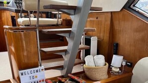 A yacht's cabin with a wooden console, a 'WELCOME TO OHANA' sign, a cushioned seating area, and a small table with a lamp and a basket of items. - Superyacht on 5* Town Quay Marina, Southampton (Southampton)