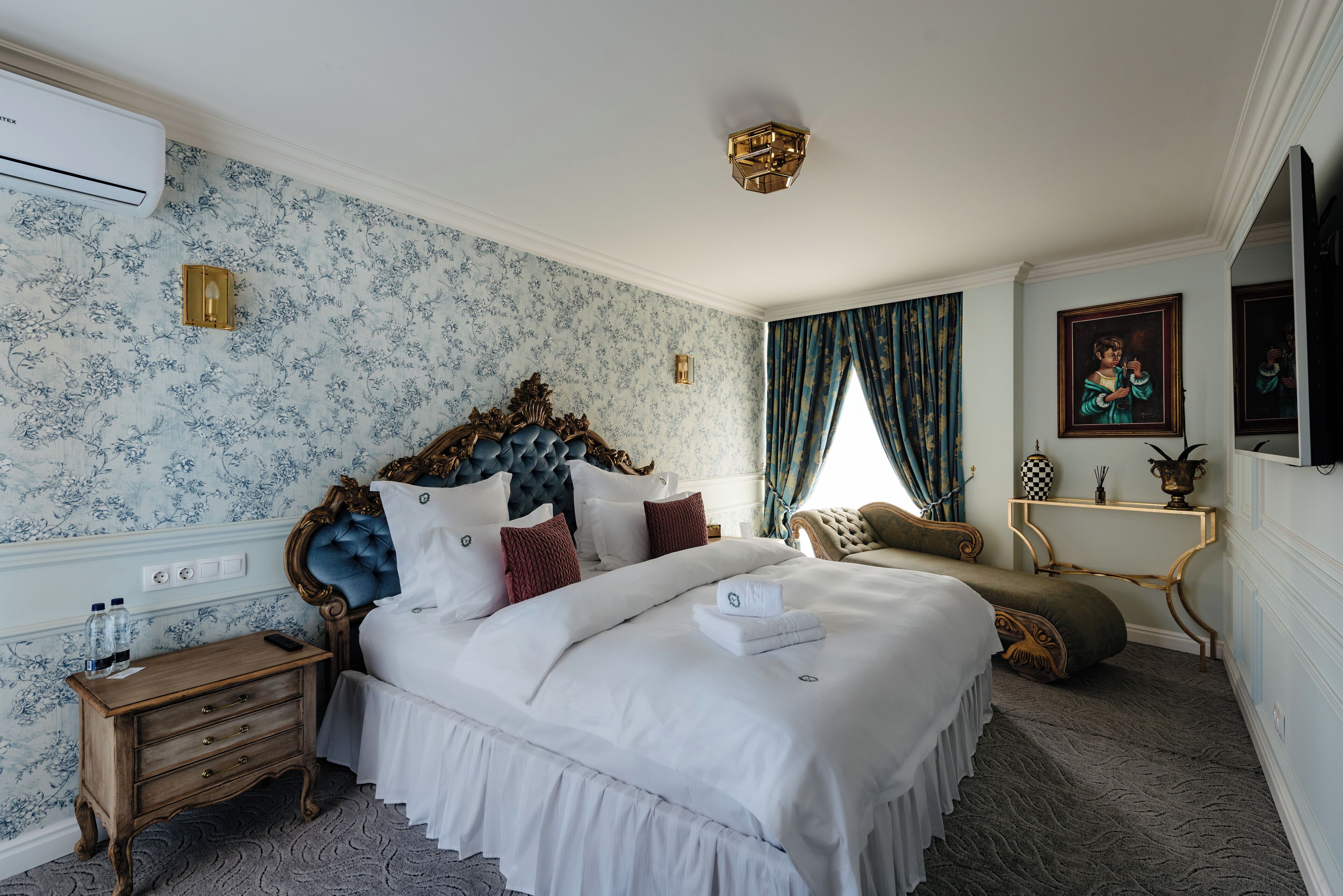 A neatly made bed with white linens, a wooden nightstand with a remote control, and a wall-mounted air conditioner.