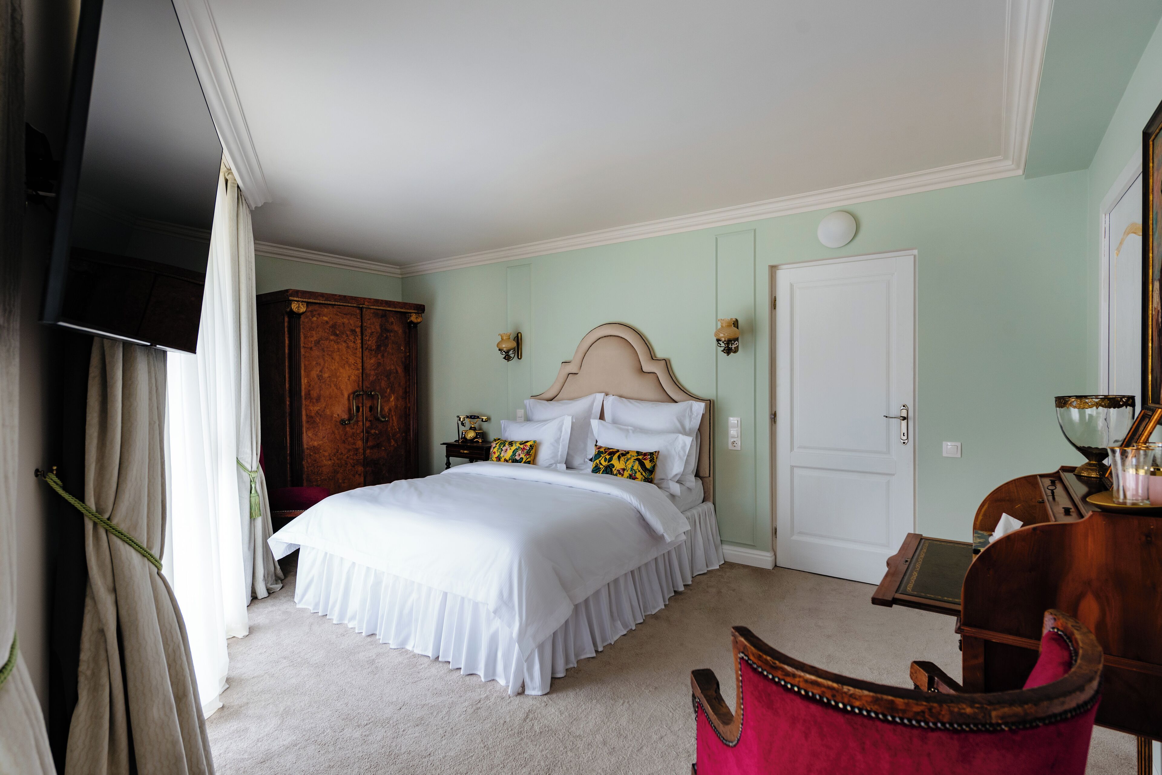 A bedroom with a large bed, a wooden wardrobe, a desk with a chair, and a door.