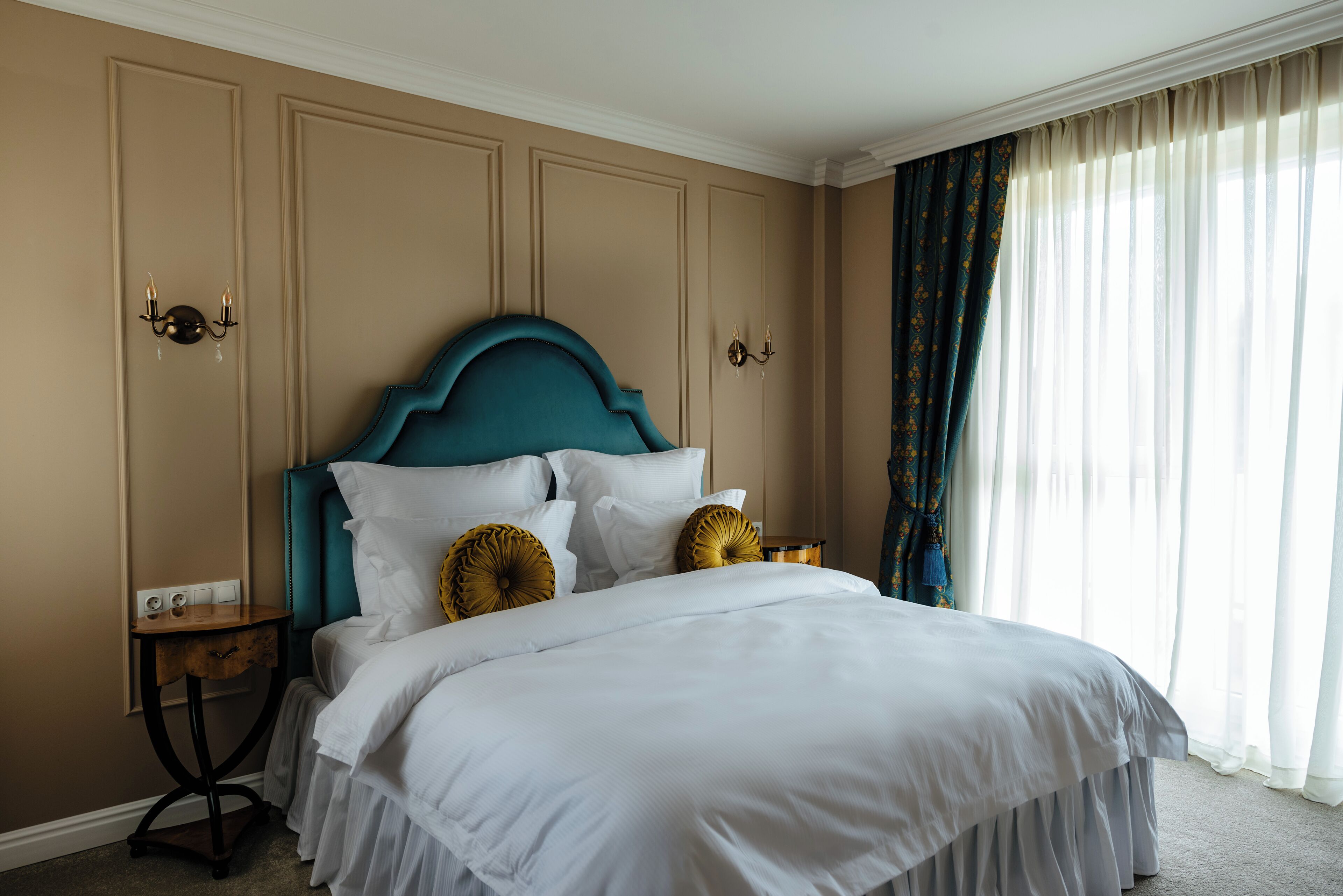 Una cama bien hecha con ropa de cama blanca y un cabecero color turquesa, una mesita de noche de madera con cajón y una lámpara fijada a la pared.