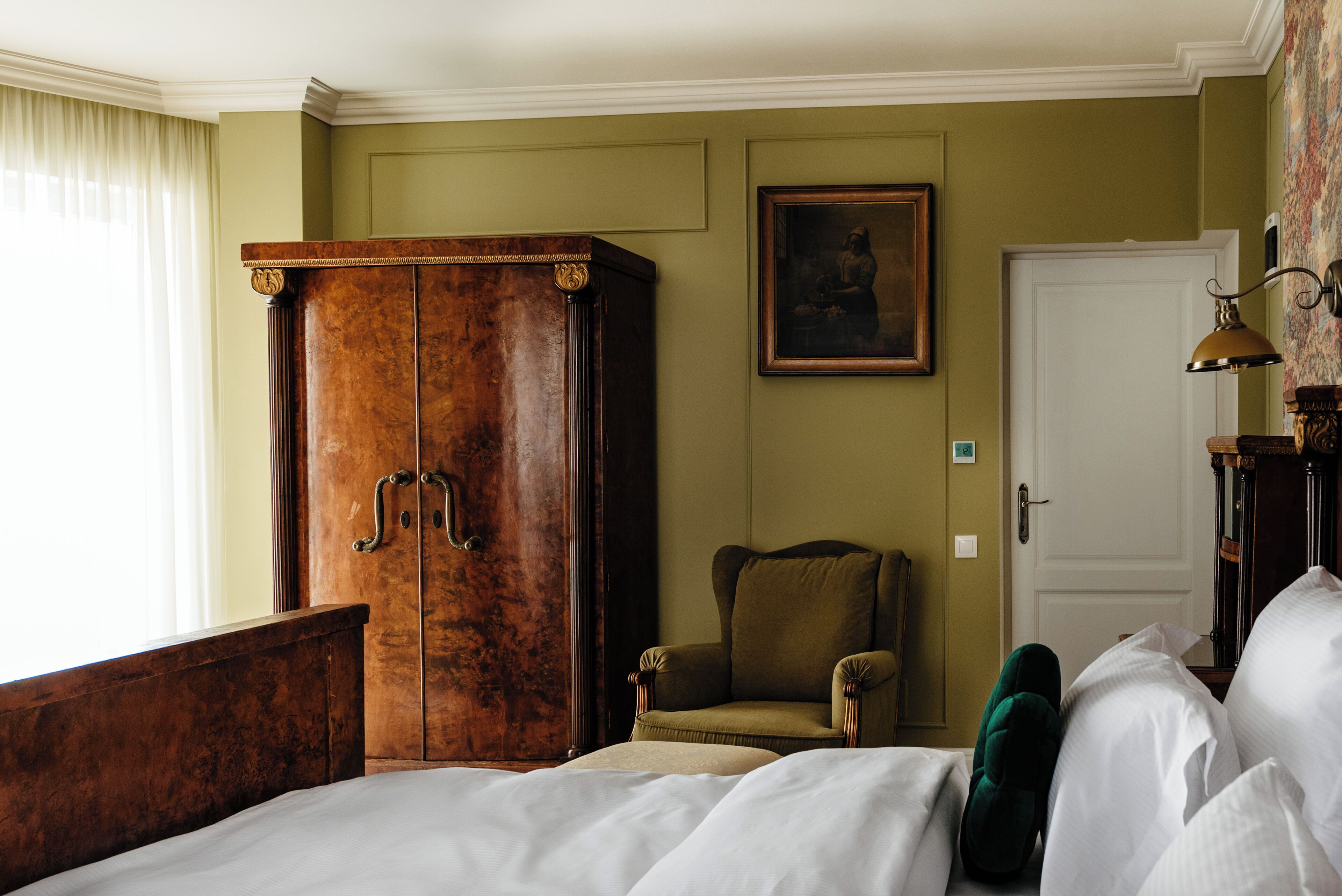 A hotel room with a large wooden wardrobe, a green armchair, a bed with white linens, and a painting on the wall.