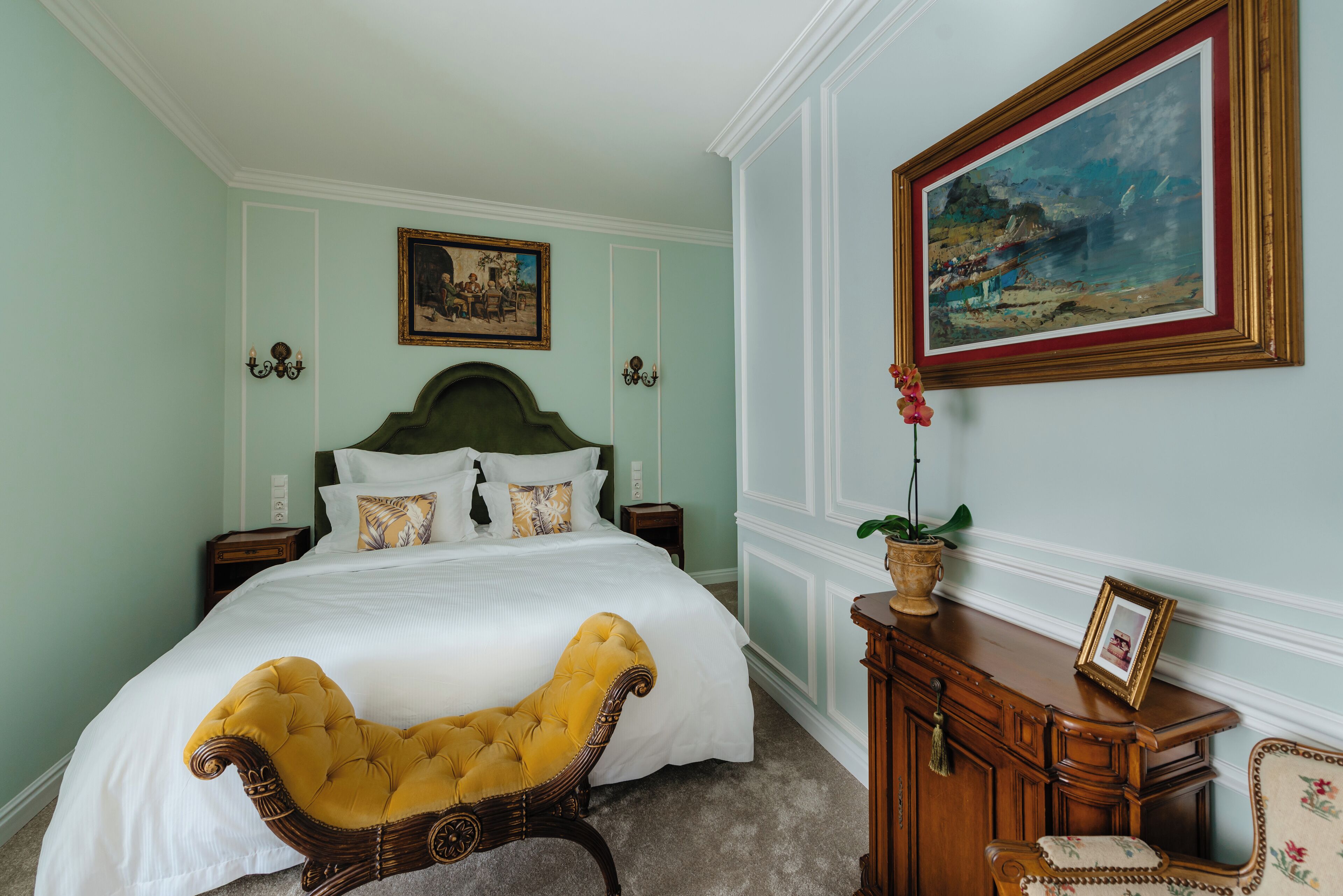 Um quarto bem organizado com uma cama grande, uma mesinha de cabeceira de madeira, uma poltrona amarela estofada, um armário e um quadro emoldurado na parede.