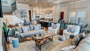 A spacious living room with a large sofa set, a coffee table, and a staircase leading to a kitchen area. - Eastern Lake Beach Access, Heated Pool, 6 Seater Golf Cart!! (Santa Rosa Beach)