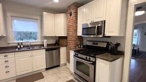 A modern kitchen with white cabinets, stainless steel appliances, a brick accent wall, and a window providing a view of greenery. - Loung Room Retreat - 3 Blocks to Mayo Campus (Rochester)