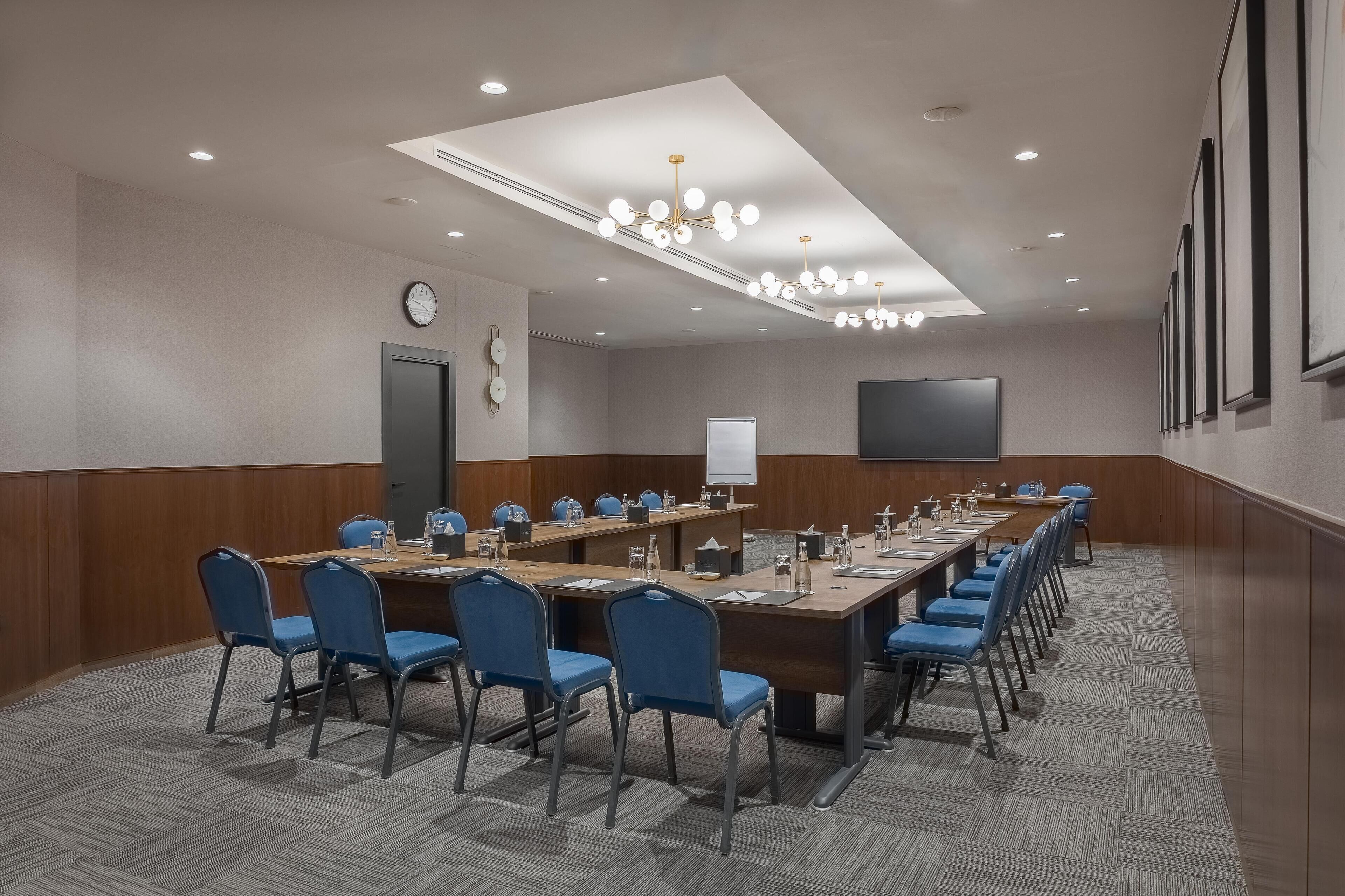 A conference room with a long table, blue chairs, a clock, and a projector screen.