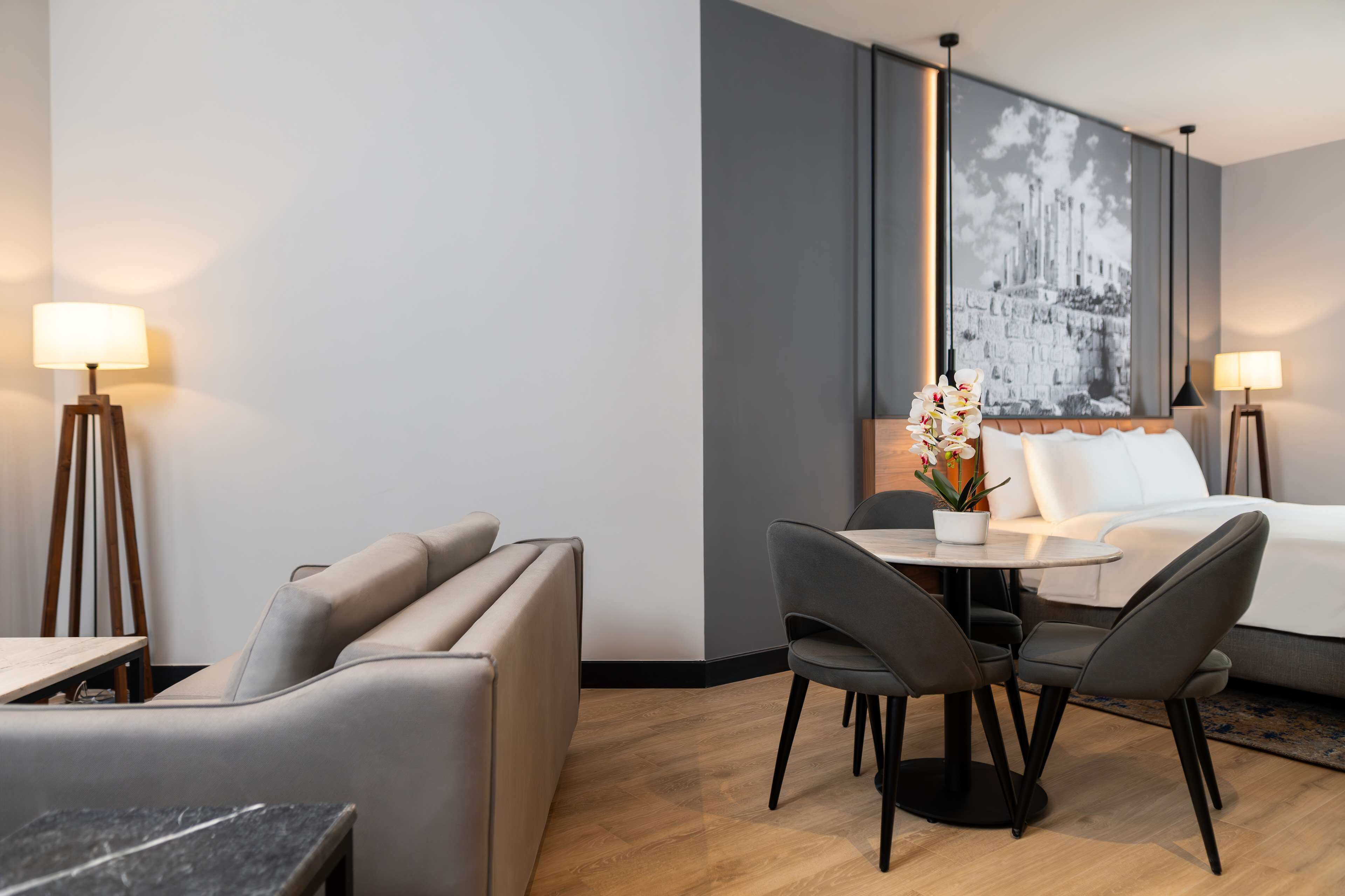A modern hotel room with a bed, a dining area with a table and chairs, and a large framed picture on the wall.