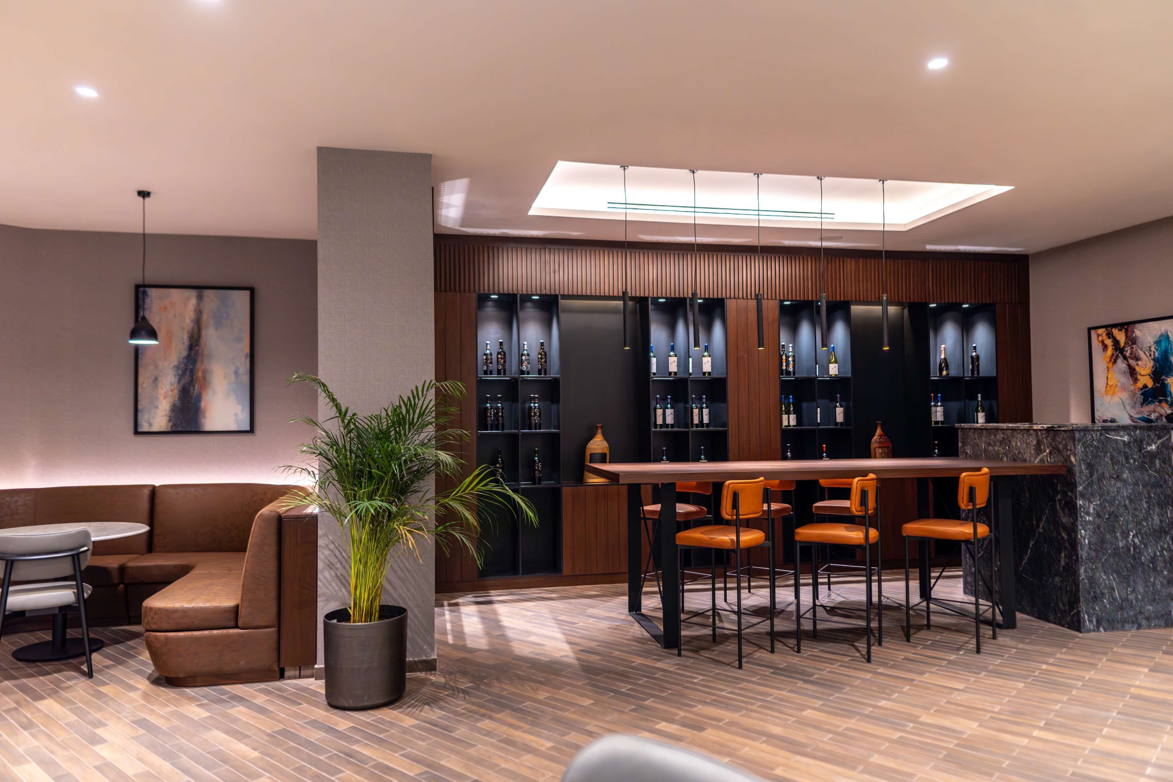 A modern bar area with a wooden bar counter, multiple shelves of bottles, and a seating arrangement with chairs.