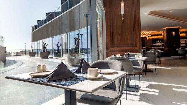 A modern outdoor dining area with tables, chairs, and a laptop. There are large windows offering a view of the cityscape. - Radisson Blu Hotel, Amman Galleria Mall (Amman)