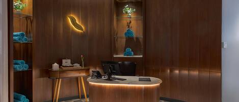 A reception area with a curved wooden reception desk, a check-in counter, and a seating area with chairs. There are shelves with towels and a potted plant.