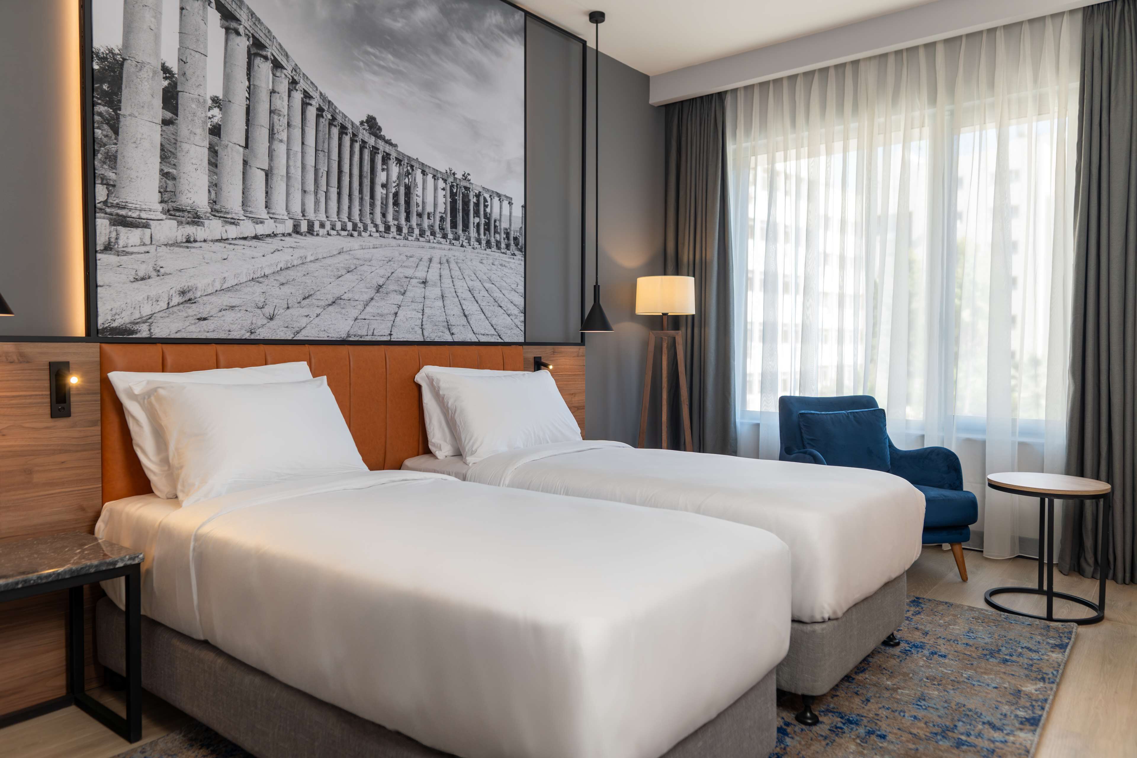 A hotel room with two beds, a large window with curtains, a blue chair, a small table, and a wall-mounted lamp.