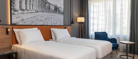 A hotel room with two beds, a large window with curtains, a blue chair, a small table, and a wall-mounted lamp.