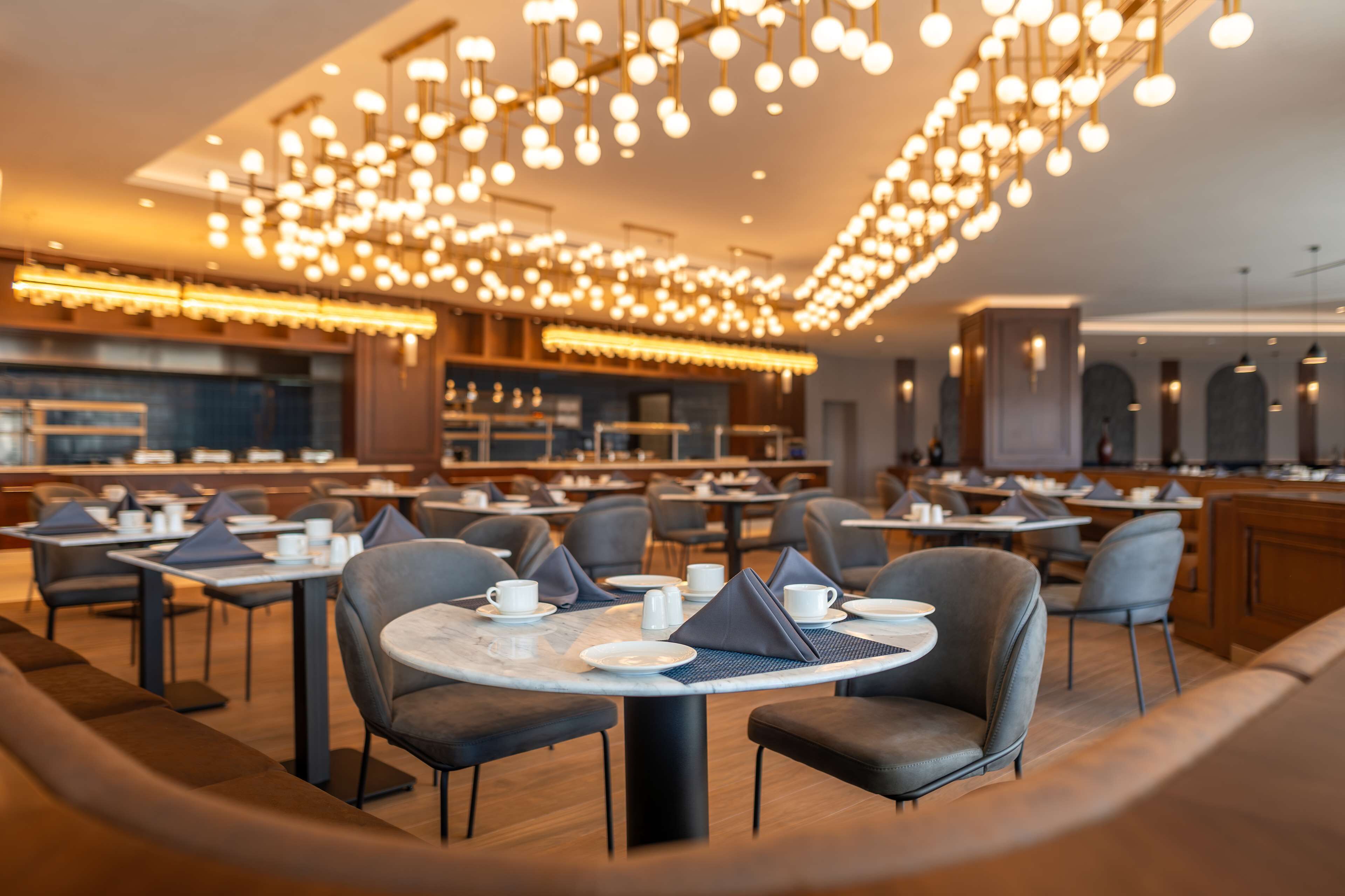 Un restaurante moderno con mesas redondas, sillas grises y un techo iluminado y cálido.