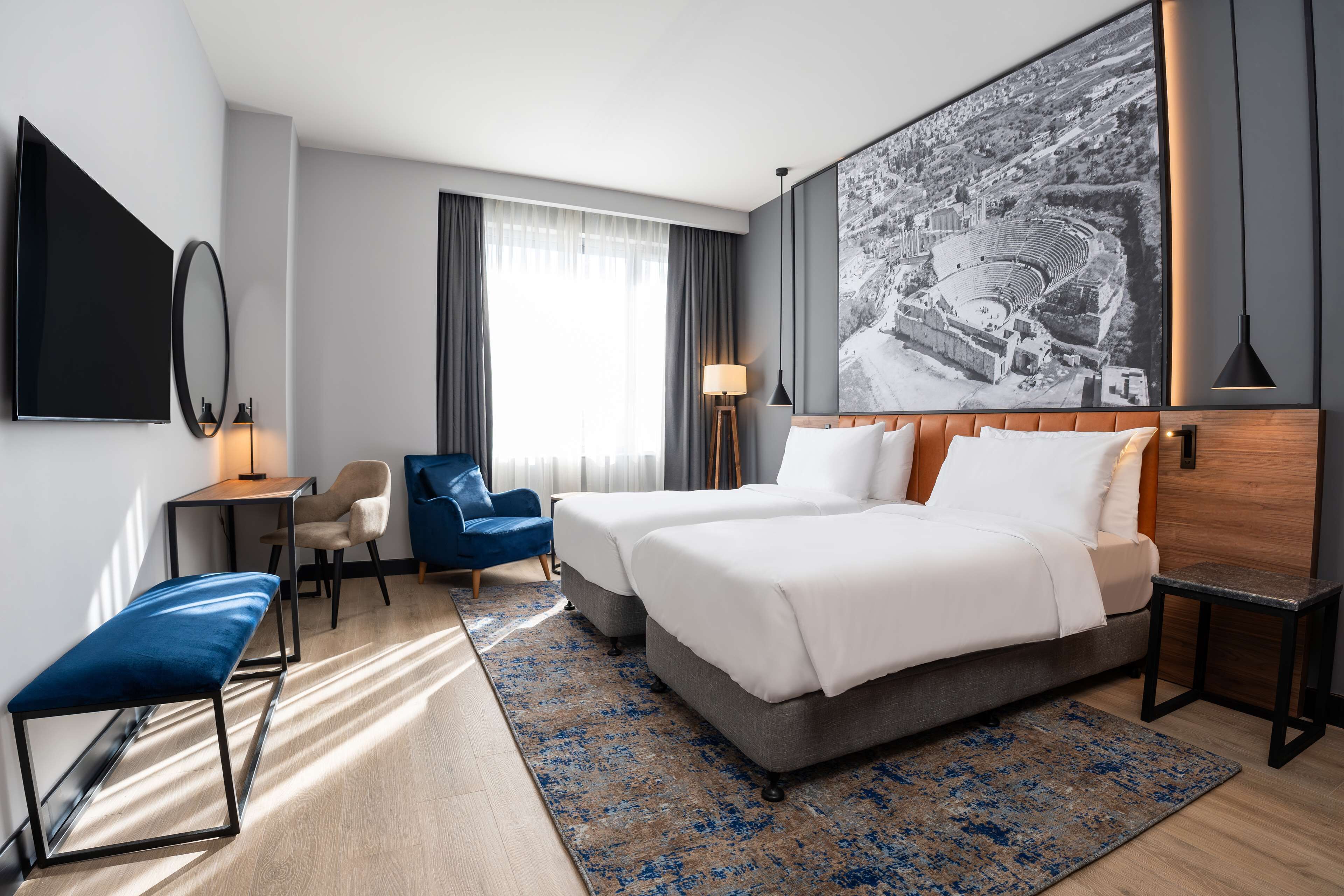 A hotel room with two beds, a desk, a chair, a bench, and a large wall-mounted TV.