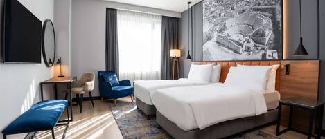A hotel room with two beds, a desk, a chair, a bench, and a large wall-mounted TV.