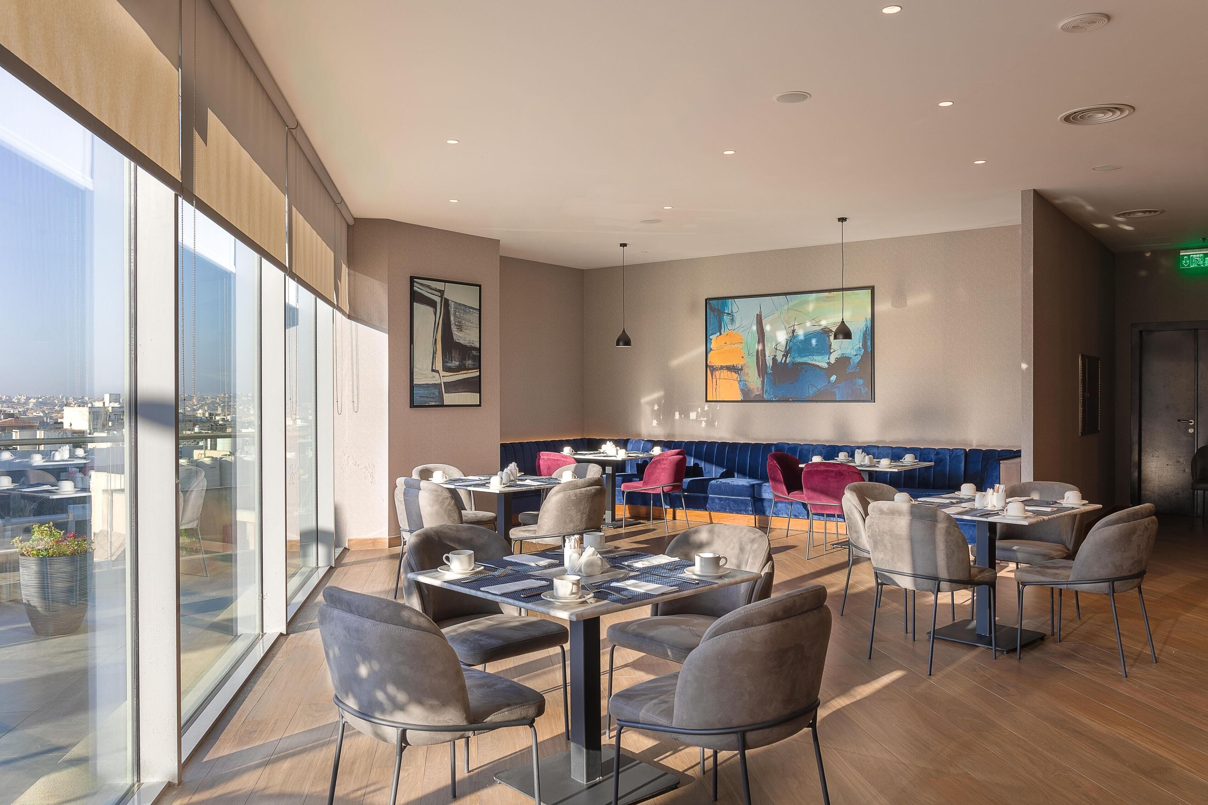 Un restaurante moderno con techo alto, ventanas amplias y una variedad de zonas de estar, incluyendo mesas y un asiento tipo banca azul.