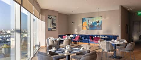 Un restaurante moderno con techo alto, ventanas amplias y una variedad de zonas de estar, incluyendo mesas y un asiento tipo banca azul.
