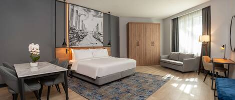 A modern hotel room with a bed, a desk, a sofa, and a dining area.